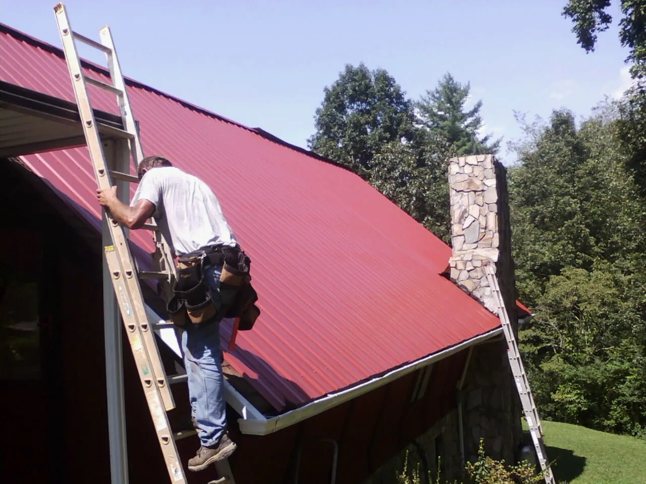 Roofer on a ladder beside a red metal roof and stone chimney — Soffit & Fascia Repair by Roofing Brothers in Elizabeth